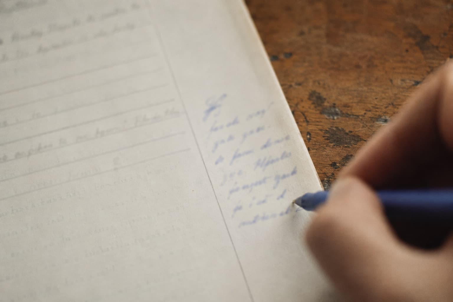 Close-up of a teacher's hand writing a comment in blue pen in the margin of a student's lined work, on a worn wooden desk